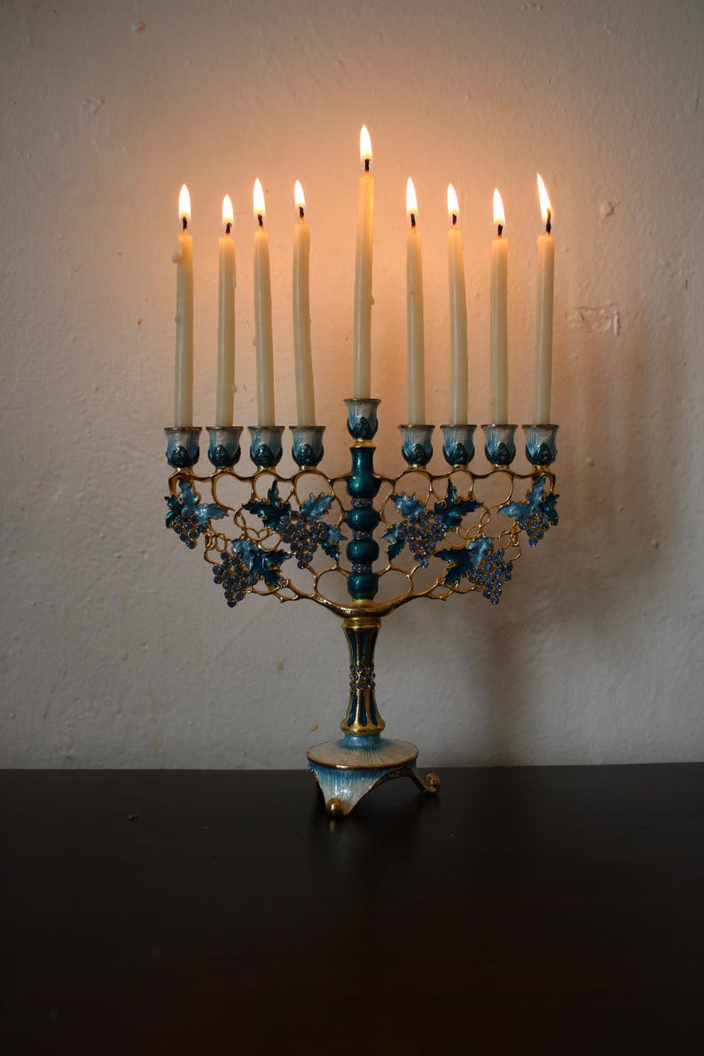 May include: A lit menorah with nine candles. The candle holders are teal and gold, with blue grape vine designs. The base is teal and gold. The candles are lit, with flames at the top.