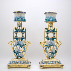 May include: Pair of ornate, gold-colored candle holders with blue and white enamel accents. Each holder features a Star of David design, floral motifs, and a square base. The candle holders are approximately 8 inches tall.