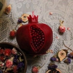 May include: A deep red pomegranate-shaped candle with a detailed seed cavity. The candle sits on a lace tablecloth, surrounded by dried flowers and figs, creating a rustic, decorative display.