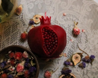 May include: A deep red pomegranate-shaped candle with a detailed seed cavity. The candle sits on a lace tablecloth, surrounded by dried flowers and figs, creating a rustic, decorative display.