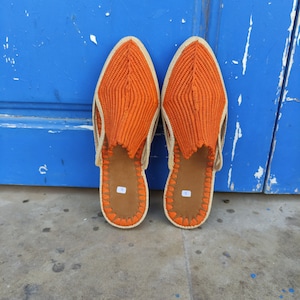 May include: A pair of orange woven leather slippers with a pointed toe and a beige trim. The slippers are sitting on a concrete surface in front of a blue door.