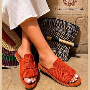 May include: Orange woven sandals with a tassel detail. The sandals are handmade and feature a flat sole. The image also shows a woven bag and the text "Essaouirahandmade".