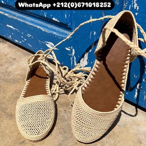May include: A pair of beige woven sandals with brown insoles and ankle ties. The sandals have a flat sole and a textured design. The background is a weathered blue surface.