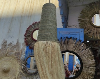 May include: A large, woven, cone-shaped lampshade with a fringe of natural fibers hangs from the ceiling. The lampshade is made of a natural material, possibly straw or rope. The lampshade is hanging in front of a blue wall with a mirror and other decorative items.
