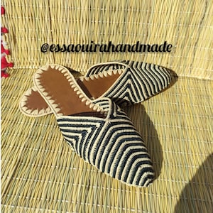 May include: A pair of black and white striped woven slippers with brown leather soles. The slippers are on a woven mat. The text '@essaouirahandmade' is visible in the image.