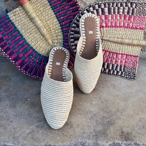 May include: Two pairs of beige woven straw slippers with a pointed toe. The slippers are sitting on a light brown surface.