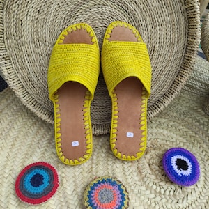 May include: A pair of yellow woven leather sandals with a brown leather sole. The sandals are on a woven straw mat with colorful crocheted circles.