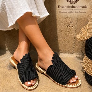 May include: Black woven slide sandals with a tassel detail. The sandals have a brown sole and a cream-coloured trim. The sandals are handmade and the text "Essaouirahandmade" is visible.