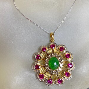 Può includere: Una collana con un ciondolo in oro con una pietra preziosa verde circondata da pietre preziose rosa e diamanti. Il ciondolo ha la forma di un fiore.