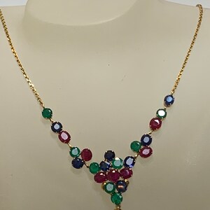 Puede incluir: Un collar de cadena de oro con un racimo de piedras preciosas rojas, verdes y azules. Las piedras preciosas están dispuestas en forma de cascada, con la piedra más grande en la parte inferior.