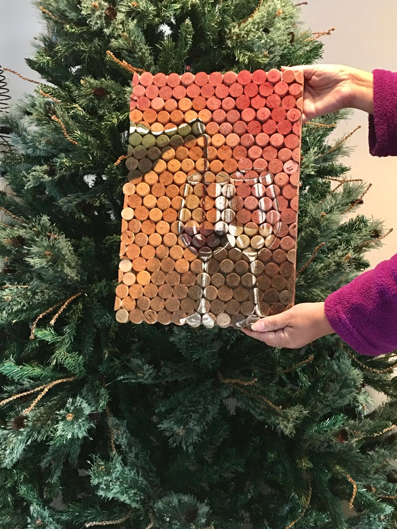 Puede incluir: Una pieza de arte de pared hecha de tapones de vino dispuestos en un patr&oacute;n de cuadr&iacute;cula. Los tapones son en tonos de marr&oacute;n, naranja y rojo. La imagen representa dos copas de vino con una botella de vino en el fondo.