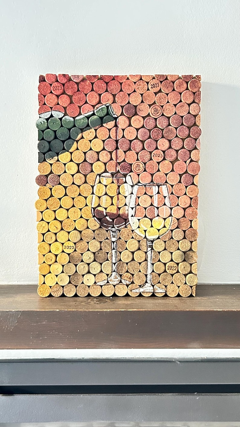 Puede incluir: Una obra de arte de pared hecha de tapones de vino, con dos copas de vino y una botella de vino. Los tapones est&aacute;n dispuestos en un patr&oacute;n de cuadr&iacute;cula y tienen diferentes tonos de marr&oacute;n y rojo. Los a&ntilde;os 2022, 2023 y 2025 est&aacute;n impresos en algunos de los tapones.
