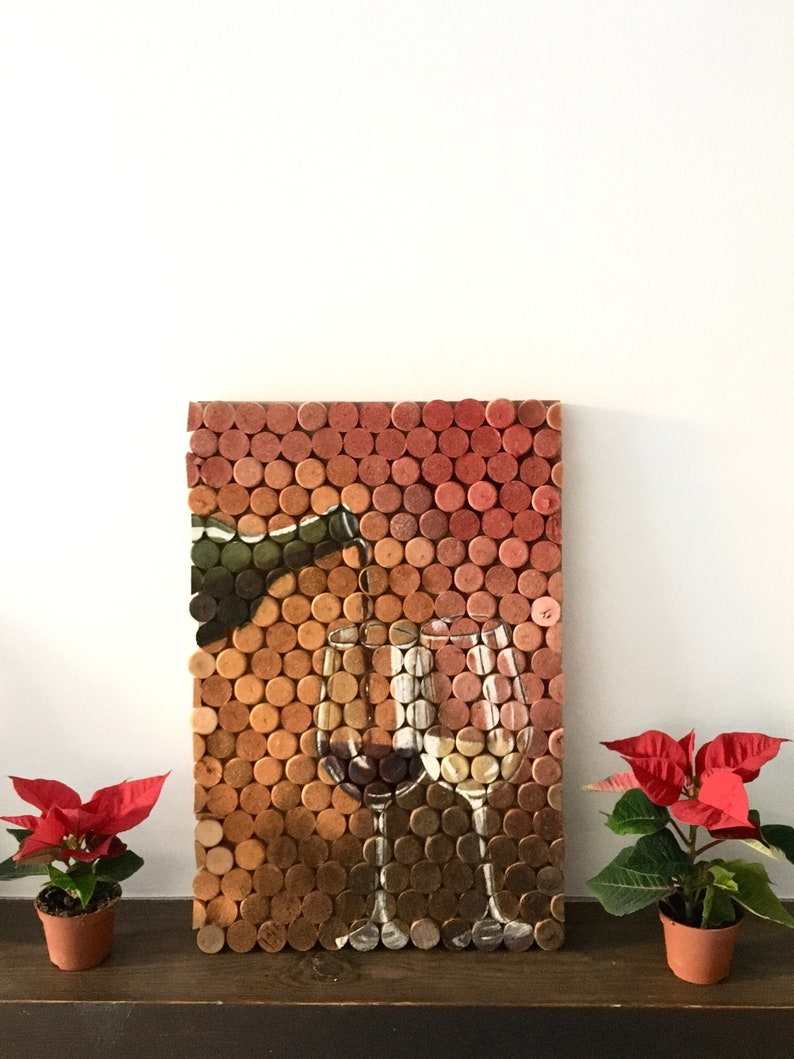 Puede incluir: Una pieza de arte de pared hecha de corchos de vino, que representa una botella de vino vertiendo en dos copas de vino. Los corchos est&aacute;n dispuestos en un patr&oacute;n de cuadr&iacute;cula y son de varios tonos de marr&oacute;n y rojo.