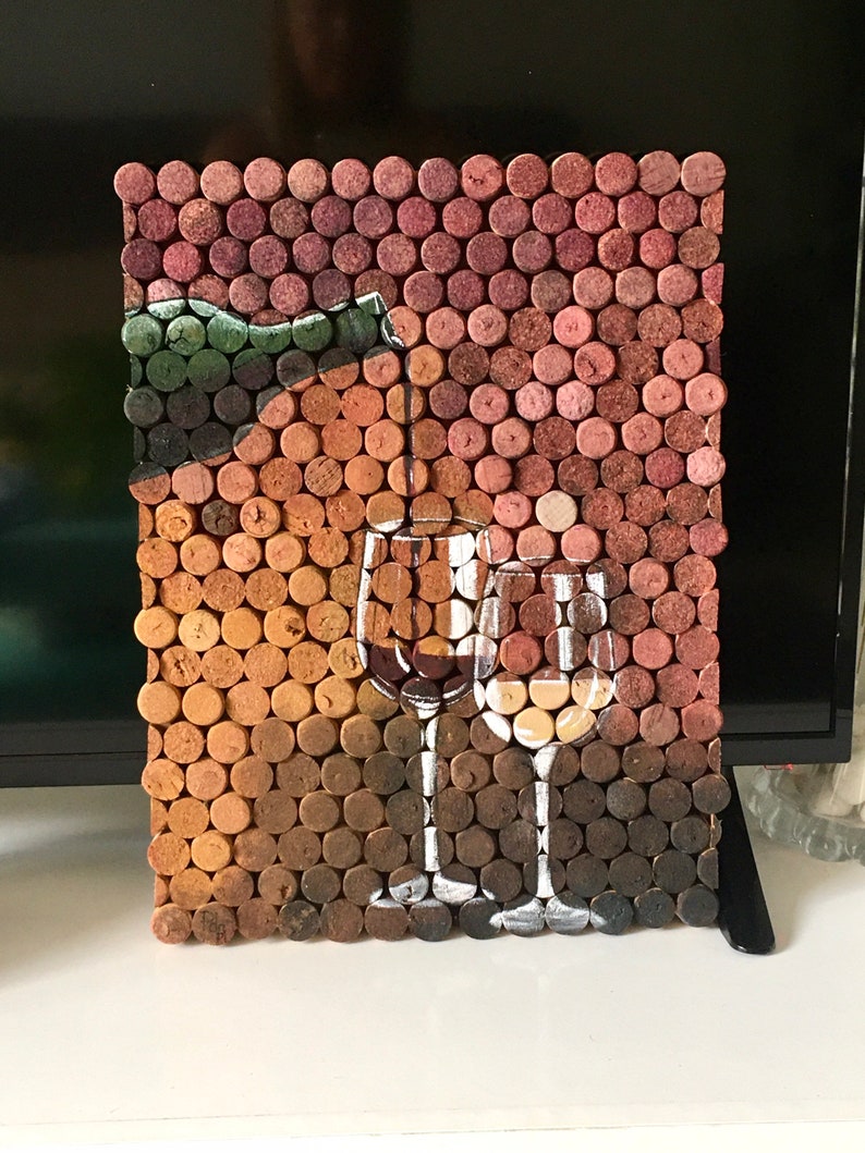 Puede incluir: Una pieza de arte de pared hecha de tapones de vino, que representa una botella de vino y dos vasos. Los tapones est&aacute;n dispuestos en un patr&oacute;n de cuadr&iacute;cula y pintados con pintura marr&oacute;n, roja y blanca.
