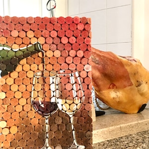 Puede incluir: Un tablero de corcho con un mosaico de tapones de vino, que presenta una imagen pintada de dos copas de vino con vino tinto y blanco. El tablero de corcho est&aacute; colgado en una pared junto a una gran pieza de jam&oacute;n curado.
