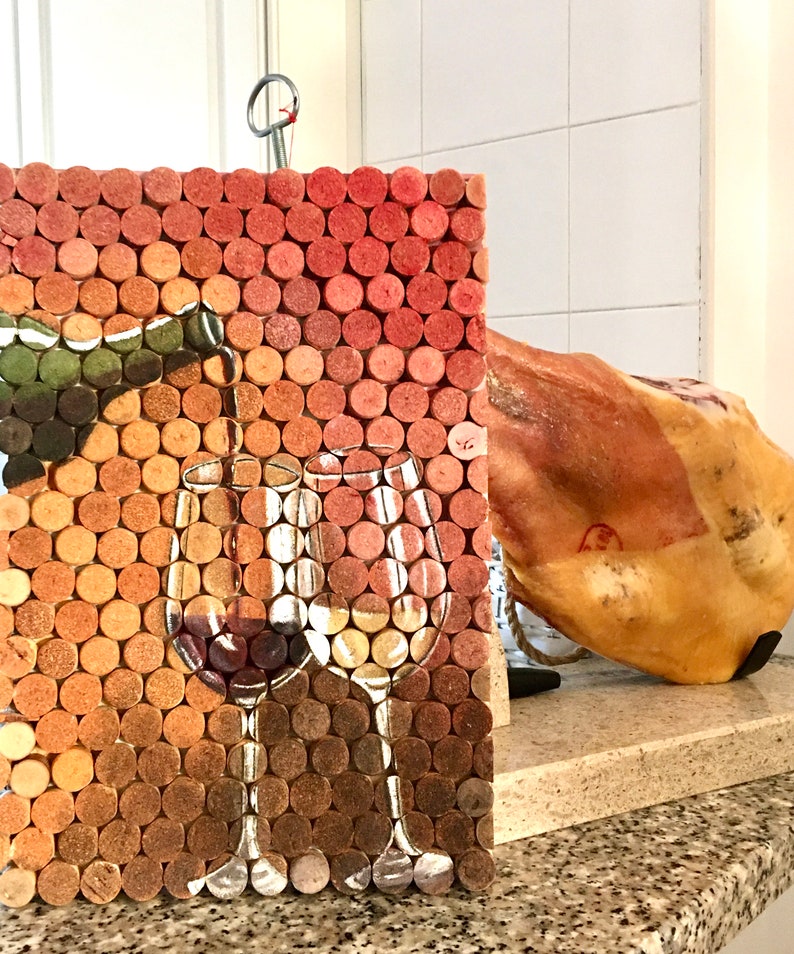Puede incluir: Un tablero de corcho con un mosaico de tapones de vino, que presenta una imagen pintada de dos copas de vino con vino tinto y blanco. El tablero de corcho est&aacute; colgado en una pared junto a una gran pieza de jam&oacute;n curado.