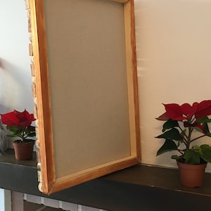 Puede incluir: Un marco de fotos de madera con un acabado de madera marr&oacute;n claro. El marco est&aacute; abierto y apoyado en una superficie marr&oacute;n oscura. Hay dos peque&ntilde;as plantas en maceta con flores rojas en el fondo.