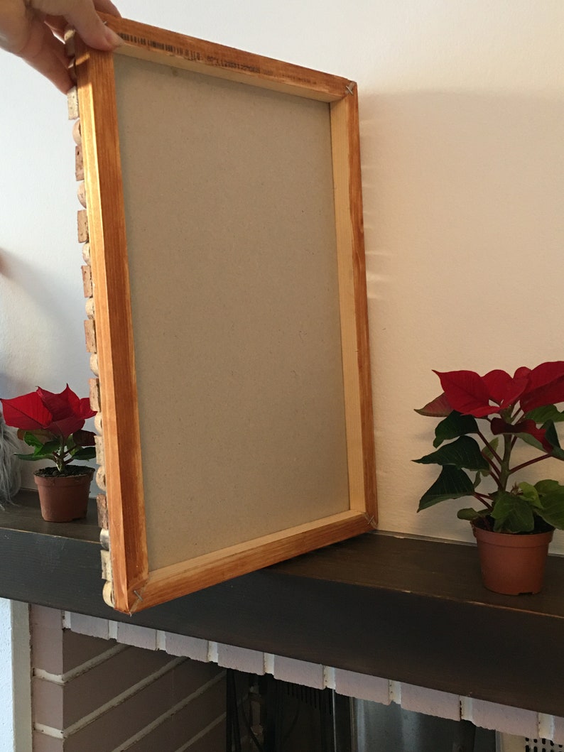 Puede incluir: Un marco de fotos de madera con un acabado de madera marr&oacute;n claro. El marco est&aacute; abierto y apoyado en una superficie marr&oacute;n oscura. Hay dos peque&ntilde;as plantas en maceta con flores rojas en el fondo.