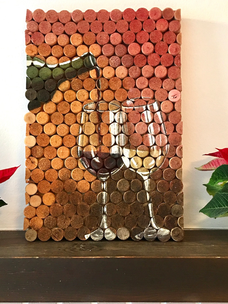 Puede incluir: Una obra de arte de pared hecha de tapones de vino, que representa dos copas de vino y una botella de vino. Los tapones est&aacute;n dispuestos en un patr&oacute;n de cuadr&iacute;cula y pintados para crear la imagen. El fondo es una pared blanca.