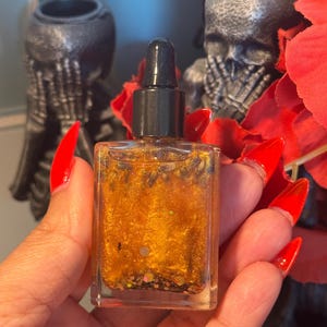 Puede incluir: Una pequeña botella de vidrio con un gotero negro lleno de un líquido dorado y purpurina. La botella está sentada en una mano.