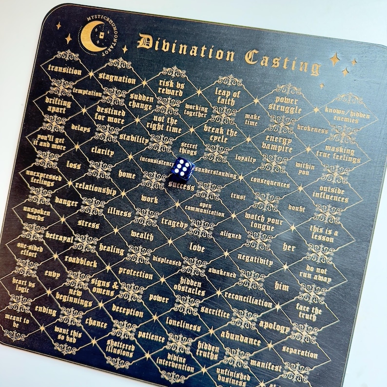 May include: A black wooden divination board with gold lettering and a blue die. The board is divided into a grid with words like "transition", "stagnation", "risk vs reward", "leap of faith", "power struggle", "energy vampire", "love", "hidden obstacles", "sacrifice", "abundance", and "separation".
