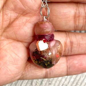 Può includere: Una collana con un ciondolo a forma di bottiglia di vetro a forma di cuore e una catena d'argento. La bottiglia è piena di fiori secchi, erbe e glitter. Un piccolo ciondolo a forma di cuore bianco è appeso alla bottiglia.
