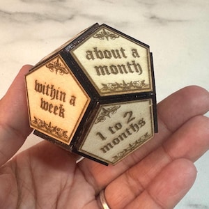 Puede incluir: Un dodecaedro de madera con texto grabado con láser que indica plazos: "within a week", "about a month" y "1 to 2 months". El objeto tiene un marco oscuro y paneles de color claro con adornos decorativos.