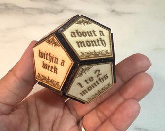 May include: A wooden dodecahedron with laser-etched text indicating timeframes: "within a week," "about a month," and "1 to 2 months." The object has a dark frame and light-colored panels with decorative flourishes.