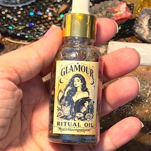 May include: A clear glass dropper bottle containing ritual oil with a gold-coloured cap. The label features a cream background with black text reading GLAMOUR RITUAL OIL Mysticbluumoontarot, and an illustration of a person with long hair.