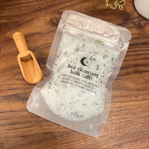 Peut inclure: Un sachet en plastique transparent rempli de sels de bain blancs et d'herbes vertes. Le sachet porte une étiquette noire avec le texte "hez cleansing bath salts" et une liste d'avantages : "curse breaking, evil removal, negative energy removal, uncrossing, psychic defense".