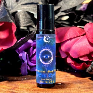 May include: A small, dark blue glass bottle with a black roll-on top. The bottle has a label with a crescent moon and an eye design. The label reads "Protection Good Luck Evil Eye" and "Sold as Curio Net Wt 24 g".