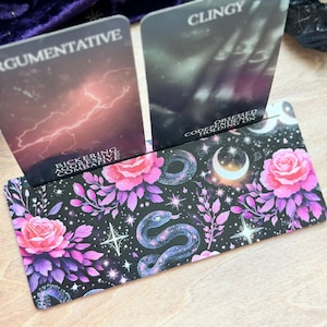 Pode incluir: Um porta-cartões com duas cartas de tarô. O suporte tem um fundo preto com rosas cor-de-rosa, folhas roxas, uma cobra e luas crescentes. As cartas dizem "ARGUMENTATIVE" e "CLINGY".