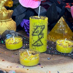 May include: Four yellow candles with black markings. The larger candle has a sigil carved into it. The candles are on a wooden surface with black and gold glitter.