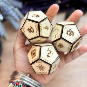 Puede incluir: Tres dados crema de múltiples caras con intrincadas ilustraciones en línea negra de símbolos y diseños botánicos. Los dados se sostienen en una mano, mostrando su detallada artesanía y estética única.