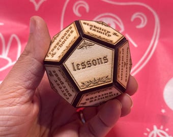 May include: A wooden dodecahedron with engraved text on each face, including "lessons," "stability & family," and "career & purpose." The edges are dark, and the background is pink with a repeating pattern.