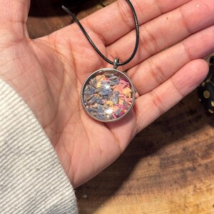 以下が含まれることがあります： 乾燥花とハーブが入った丸いガラス製のチャームがついたシルバーのペンダントネックレス。チャームは黒い紐で吊り下げられています。