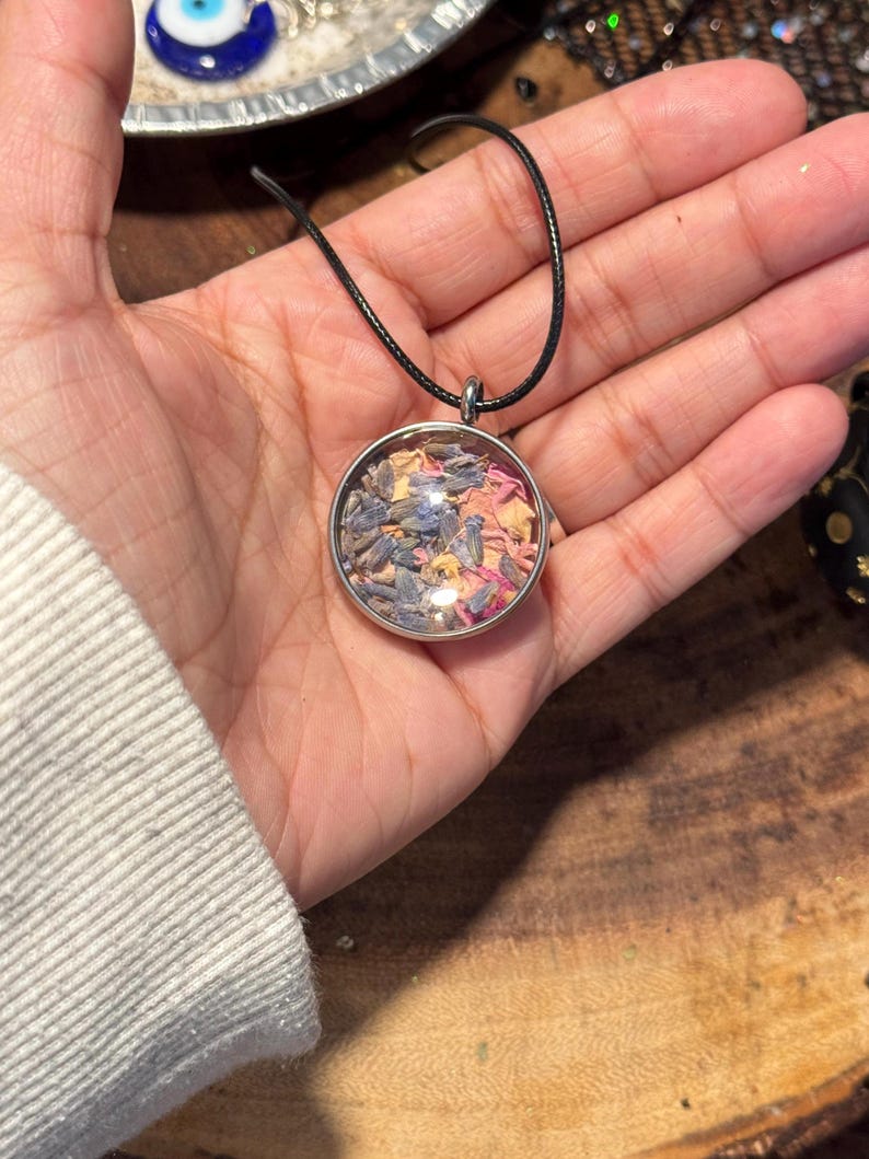以下が含まれることがあります： 乾燥花とハーブが入った丸いガラス製のチャームがついたシルバーのペンダントネックレス。チャームは黒い紐で吊り下げられています。