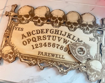 Puede incluir: Un tablero de Ouija de madera con una planchette y un diseño de calavera. El tablero presenta las letras A-Z, los números 1-9 y las palabras "YES", "NO" y "FAREWELL".