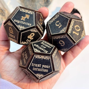 Puede incluir: Tres dados dodecaédricos negros con letras y detalles dorados. Cada dado presenta símbolos astrológicos, números y frases como "trust your intuition" y "blessings are coming". Los dados se sostienen en una mano.