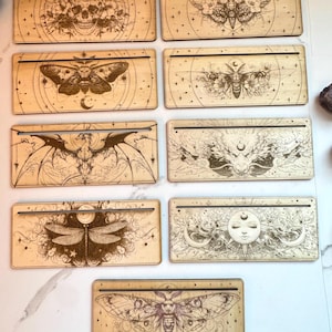 May include: Eight wooden rectangular signs with intricate laser-cut designs featuring moths, skulls, and celestial imagery. The signs have a light wood finish and are perfect for displaying on a shelf or wall.