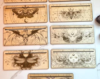 May include: Eight wooden rectangular signs with intricate laser-cut designs featuring moths, skulls, and celestial imagery. The signs have a light wood finish and are perfect for displaying on a shelf or wall.