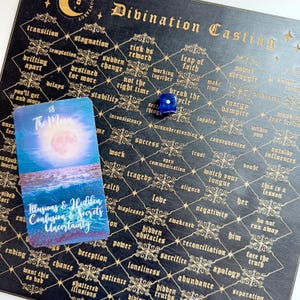 May include: A black divination board with gold accents and a blue die. The board is filled with words and phrases related to divination and fortune telling. A tarot card with the number 18 and the name "The Moon" is on the board. The card shows a blue and pink moon over a body of water.