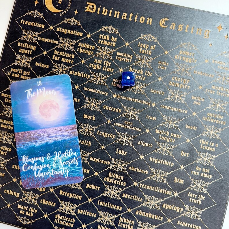May include: A black divination board with gold accents and a blue die. The board is filled with words and phrases related to divination and fortune telling. A tarot card with the number 18 and the name "The Moon" is on the board. The card shows a blue and pink moon over a body of water.