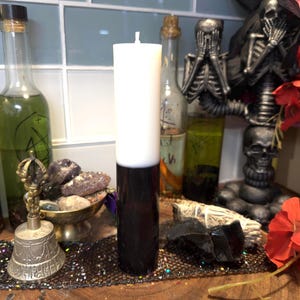 May include: A white and black pillar candle with a black base and a white top. The candle is on a table with other items, including a bell, bottles, and crystals.