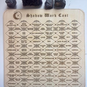 May include: A wooden board with a grid of squares, each containing a word or phrase related to shadow work. The board is titled "Shadow Work Cast".