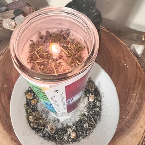 May include: A pink candle with herbs and a white label with the text "The Earth is my witness, I will not be silenced." The candle is burning and surrounded by white powder and herbs on a white plate.
