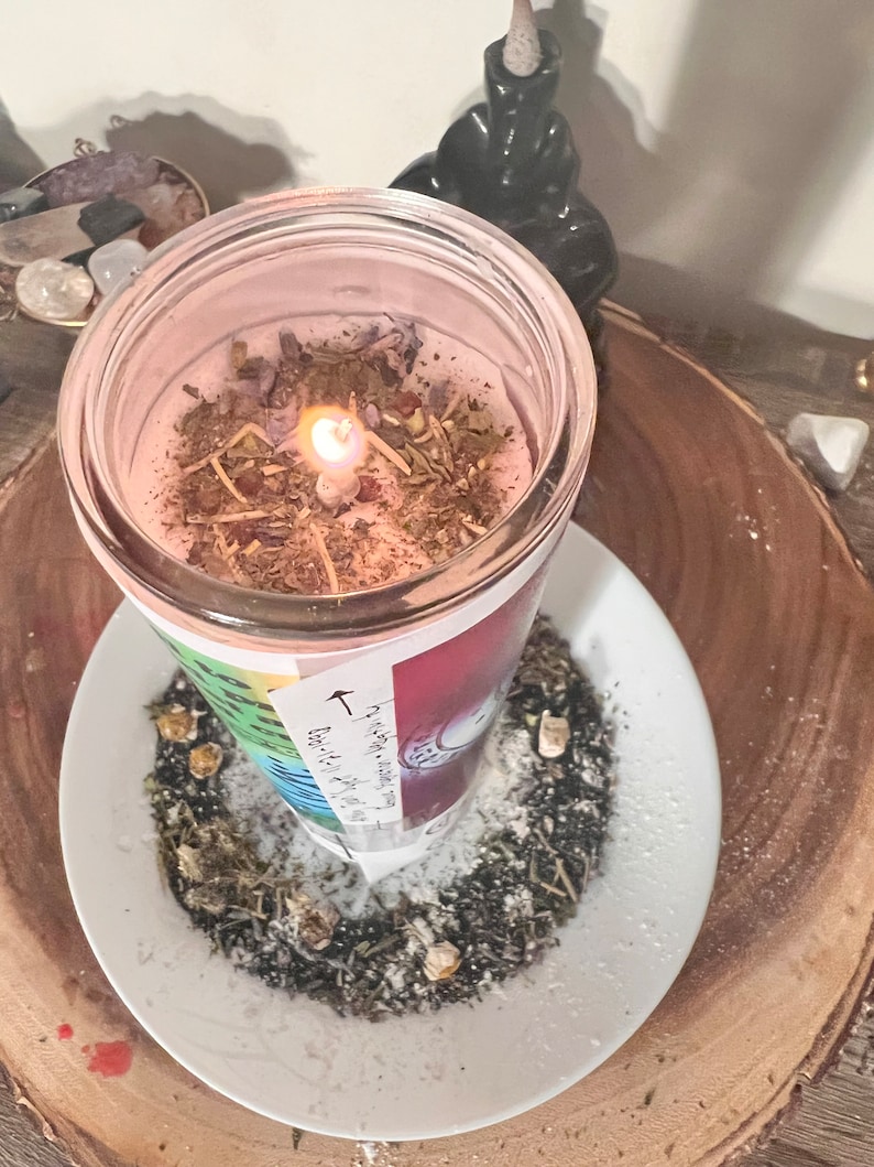 May include: A pink candle with herbs and a white label with the text "The Earth is my witness, I will not be silenced." The candle is burning and surrounded by white powder and herbs on a white plate.