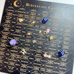Könnte beinhalten: Ein schwarzes Divinationsbrett mit goldfarbenen Buchstaben und einem Gittermuster. Das Brett zeigt verschiedene Charms und Würfel, darunter ein blauer und violetter d20-Würfel und ein blauer sechsseitiger Würfel. Der Text "Divination Casting" steht oben.