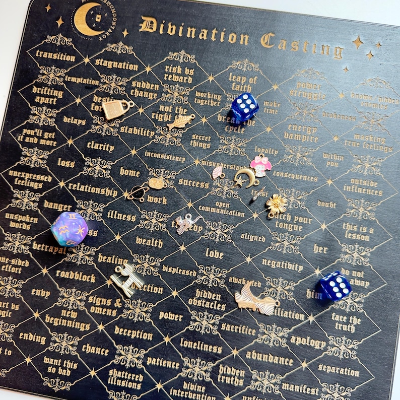 May include: A black divination board with gold lettering and a grid pattern. The board features words like "transition", "stagnation", "risk", "reward", "leap of faith", "love", "negativity", "hidden obstacles", "abundance", and "manifest". There are several charms and dice scattered on the board.