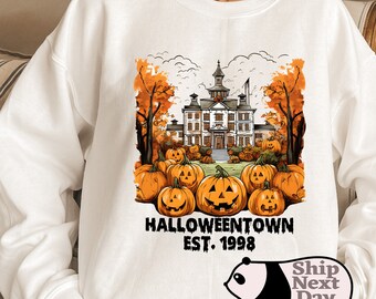 Puede incluir: Sudadera blanca con un gráfico de una casa encantada y calabazas. El texto "HALLOWEENTOWN EST. 1998" está impreso debajo del gráfico.